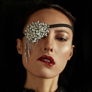 Puede incluir: Primer plano de una persona con un parche en el ojo negro adornado con perlas y cristales. Cadenas plateadas cuelgan del parche. La persona lleva pintalabios rojo y una camisa negra.