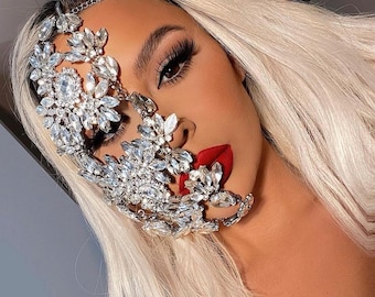 May include: A glamorous face adorned with a sparkling, ornate crystal mask. The mask, featuring floral and geometric designs, covers the eye and cheek, complemented by bold red lipstick and long, blonde hair. The mask is attached to a delicate chain that goes around the head.