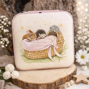 Puede incluir: Un estuche cuadrado de color rosa claro con cierre de cremallera. El estuche presenta una ilustración a la acuarela de un bebé en una cesta de mimbre, con un conejo, un erizo y un ratón. El estuche está sobre una rodaja de madera.