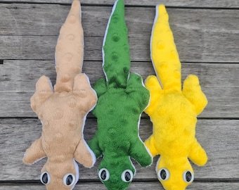 May include: Three plush toy lizards in tan, green, and yellow. Each lizard has a long tail, four small legs, and large, round eyes. The toys are made of soft, textured fabric and are lying on a wooden surface.
