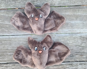 May include: Two brown plush bats with black eyes and a pink nose. The bats are lying on a wooden surface.
