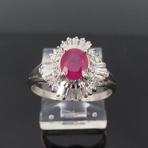 May include: A silver ring featuring a large oval ruby gemstone surrounded by smaller, clear gemstones. The ruby is a deep red color, and the ring has a classic, elegant design. The ring is displayed on a clear stand.