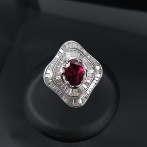 Può includere: Un anello d'argento con una grande pietra preziosa rosso scuro a taglio ovale al centro, circondata da più file di pietre preziose chiare rettangolari più piccole. L'anello ha un design geometrico unico.