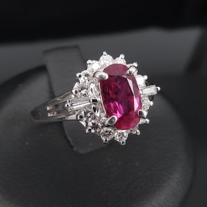 Puede incluir: Anillo de plata con un gran rubí ovalado rodeado por un halo de pequeños diamantes redondos. También presenta diamantes de talla baguette. El anillo se encuentra sobre un fondo negro.