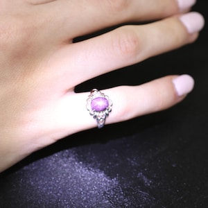 May include: A silver ring with an oval, purple gemstone. The ring has an ornate, filigree design around the gemstone. The ring is on a finger, with a dark background.
