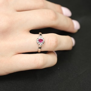 Op de afbeelding: Een zilveren ring met een ovale robijn edelsteen in het midden, omringd door kleine, heldere accentstenen. De ring wordt op een vinger getoond tegen een donkere achtergrond. De robijn is helderrood.