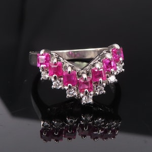 May include: A silver ring with a V-shaped design, adorned with square-cut pink gemstones and small, round white gemstones. The ring is set against a black background, creating a contrast that highlights the ring's details.
