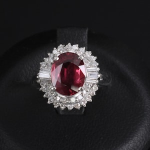 Könnte beinhalten: Ein silberfarbener Ring mit einem großen, oval geschliffenen, tiefroten Edelstein in der Mitte. Der zentrale Stein ist von einem Kranz aus kleinen, runden, klaren weißen Steinen und zwei im Baguetteschliff geschliffenen weißen Steinen umgeben.