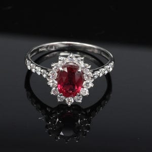 May include: A silver-toned ring featuring a central oval-cut deep red gemstone surrounded by a halo of small, clear, round-cut stones. The band is slender and adorned with additional small, clear, round-cut stones along the sides.