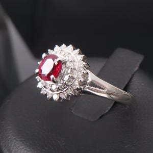 May include: A silver ring featuring a large, oval-cut ruby gemstone surrounded by a halo of smaller, clear gemstones. The ring's band is a simple, polished silver. The ruby is a deep, vibrant red.