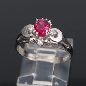 Op de afbeelding: Een zilveren ring met een grote, ovaal geslepen, helder roze edelsteen. De ring is versierd met kleinere, heldere edelstenen en decoratieve krullen. De ring wordt gepresenteerd op een transparante standaard.