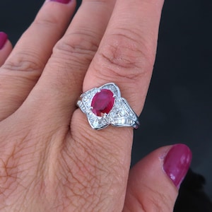 May include: A silver ring with a large oval ruby gemstone surrounded by smaller diamonds. The ring is on a person's finger.