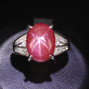 Könnte beinhalten: Ein silberfarbener Ring mit einem großen, ovalen, im Cabochon-Schliff geschliffenen roten Sternrubin als Mittelstück. Der Rubin zeigt einen deutlichen sechsstrahligen Sterneffekt. Die Ringschiene ist mit kleinen, klaren Steinen besetzt.
