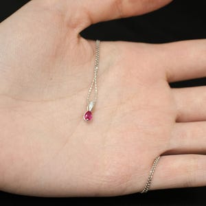 May include: A delicate silver necklace with a small, teardrop-shaped ruby pendant. The ruby is a vibrant red color and is set in a silver bezel. The necklace is shown against a neutral background.