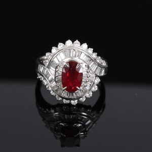 May include: A silver-toned ring featuring a central oval-cut deep red gemstone surrounded by a halo of small round diamonds and an outer layer of tapered baguette-cut diamonds arranged in a fan-like pattern on a polished metal band.