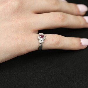 May include: A silver ring featuring a central oval ruby gemstone, surrounded by smaller, clear gemstones. The ring's band has a split design, and the ring is displayed on a hand against a black background.