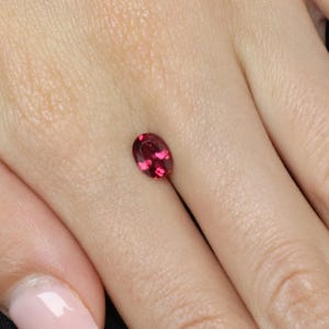 May include: A loose, oval-cut gemstone with a deep, vibrant red color and a faceted surface that reflects light. The stone is small and displays internal clarity with visible light-catching facets throughout its polished, translucent structure.