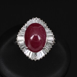 May include: A silver ring featuring a large, oval, deep red gemstone surrounded by a halo of baguette-cut, clear gemstones. The ring is set against a black background, highlighting the sparkle of the stones.