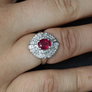 May include: A silver-toned ring featuring a central oval-cut deep red ruby gemstone. The ruby is surrounded by a double halo of small, clear, round-cut stones set in a decorative, scalloped metal frame that extends across the finger.