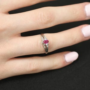 May include: A silver ring with a central oval ruby gemstone, flanked by smaller white gemstones. The ring is set on a finger, showcasing its delicate design and vibrant red and white color scheme.