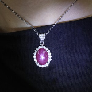 May include: A silver necklace featuring an oval-shaped, deep pink star ruby pendant surrounded by a halo of small, clear, round-cut stones. The pendant is suspended from a delicate silver chain with a polished metal bail set with tiny stones.