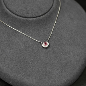 May include: A delicate silver necklace with a small pendant. The pendant features a central oval-shaped red gemstone surrounded by a halo of smaller, clear stones. The necklace is displayed on a gray velvet surface.