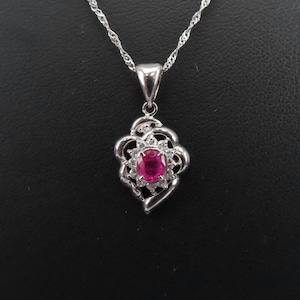 Puede incluir: Un collar con colgante de plata con un diseño floral. El colgante tiene un rubí central ovalado rodeado de pequeñas piedras preciosas transparentes. La cadena del collar es de estilo delicado y retorcido. El fondo es negro.