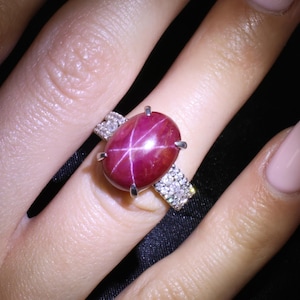 Könnte beinhalten: Ein Ring mit einem großen, ovalen, tiefroten Sternrubin-Cabochon, der einen sichtbaren sechsstrahligen Sterneffekt aufweist. Der Stein ist in eine silberfarbene Metallschiene gefasst, die mit kleinen, klaren, funkelnden Steinen besetzt ist.