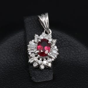 May include: A silver pendant featuring a large oval ruby gemstone at the center, surrounded by smaller diamonds and baguette-cut stones. The pendant is set on a black surface.