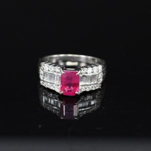 May include: A silver ring with a rectangular, vibrant red gemstone at the center, flanked by smaller, clear stones. The ring's band is also adorned with clear stones, creating a sparkling effect against the black background.