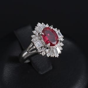 May include: A silver ring with a large, oval-cut ruby as the central stone, encircled by a halo of smaller diamonds and baguette-cut diamonds. The ring is displayed against a black backdrop, highlighting its intricate design.