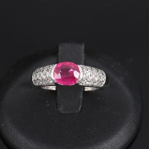 May include: A silver ring with a large, oval ruby centre stone. The band is set with numerous small diamonds. The ring is presented on a black stand, highlighting its luxurious design and the rich colour of the ruby.