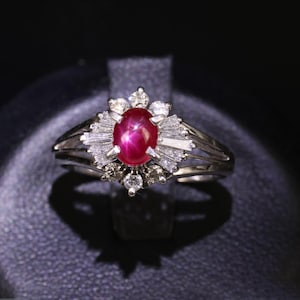May include: A silver-toned ring featuring a central oval-shaped red star ruby cabochon. The ruby is surrounded by a halo of baguette-cut and round-cut clear stones, set in a metal band that splits into two sections on each side of the setting.