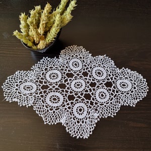 May include: A white, handmade, crocheted doily with a detailed floral pattern. The doily is placed on a dark brown surface. A small black bowl with dried green plants is in the background.