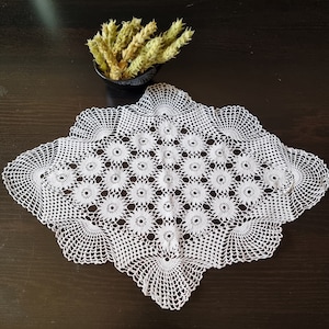 May include: White crocheted doily with a delicate floral pattern. The doily is square shaped and has a scalloped edge.