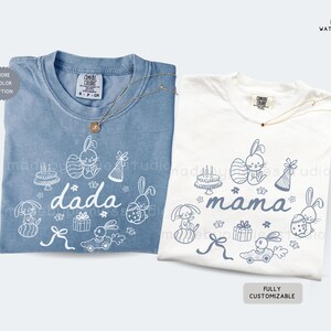 May include: Two t-shirts, one in a faded blue and the other in white, featuring bunny illustrations and text. The blue shirt says "dada", the white shirt says "mama". Both have round necks and are accessorized with a gold necklace.