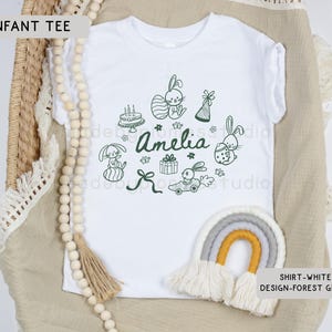 May include: White infant tee with a forest green design featuring bunnies, a cake, gifts, and the name "Amelia". The shirt is displayed on a neutral-toned surface with a decorative rainbow and wooden beads.