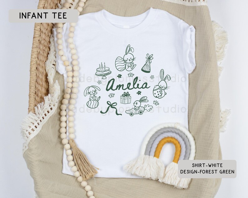May include: White infant tee with a forest green design featuring bunnies, a cake, gifts, and the name "Amelia". The shirt is displayed on a neutral-toned surface with a decorative rainbow and wooden beads.