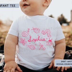 Op de afbeelding: Witte baby-t-shirt met een roze insecten- en naamontwerp. Het shirt heeft een slak, lieveheersbeestje, vlinder, libelle en rups, met de naam "Sophia" in cursief. De tekst "INFANT TEE" en "SHIRT-WHITE DESIGN-CRUNCHBERRY" zijn ook zichtbaar.