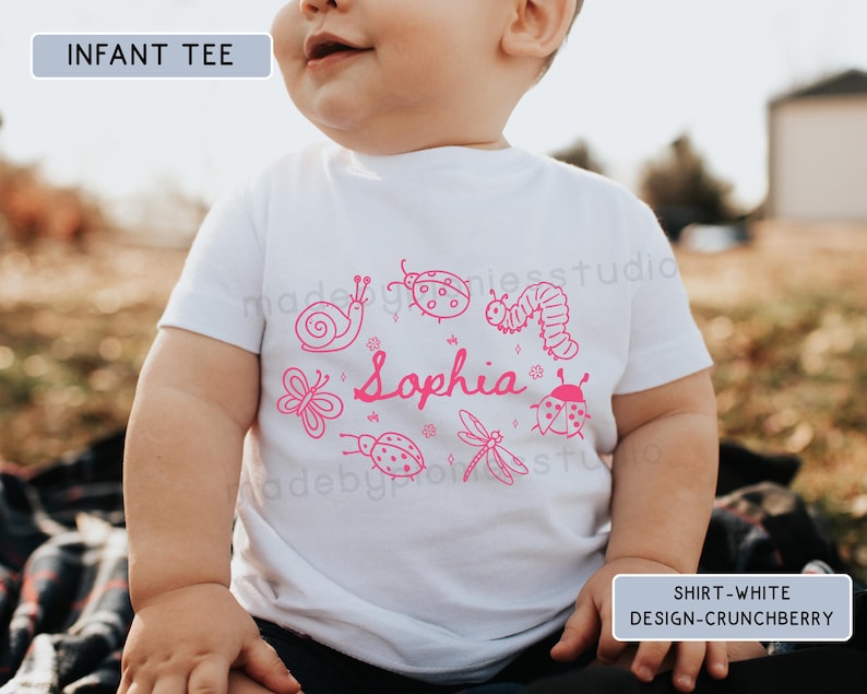 Op de afbeelding: Witte baby-t-shirt met een roze insecten- en naamontwerp. Het shirt heeft een slak, lieveheersbeestje, vlinder, libelle en rups, met de naam "Sophia" in cursief. De tekst "INFANT TEE" en "SHIRT-WHITE DESIGN-CRUNCHBERRY" zijn ook zichtbaar.