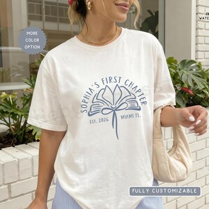 May include: A white short-sleeved t-shirt featuring a blue graphic of an open book with a ribbon tie. The text reads "SOPHIA'S FIRST CHAPTER" in an arc above the book, with "EST. 2026" and "MIAMI FL." printed below the graphic design.