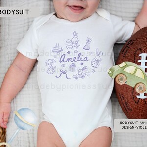 May include: White baby bodysuit featuring a violet illustration of bunnies, a cake, gifts, and a car. The name "Amelia" is written in violet script. A brown football and a green toy car are next to the bodysuit.