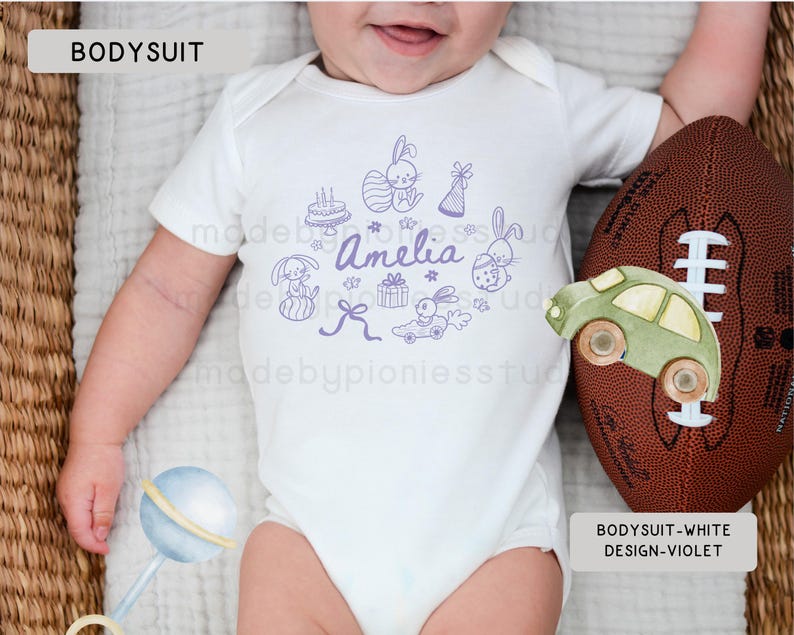 May include: White baby bodysuit featuring a violet illustration of bunnies, a cake, gifts, and a car. The name "Amelia" is written in violet script. A brown football and a green toy car are next to the bodysuit.