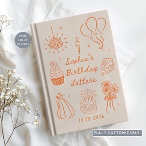 May include: A light beige hardcover book with orange line art illustrations including a disco ball, balloons, a cupcake, a slice of cake, a pumpkin, a heart-shaped cake, and flowers. The text reads Sophia's Birthday Letters and 10-10-2026.