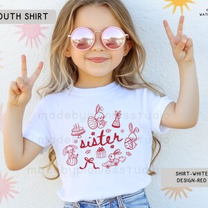 May include: White youth t-shirt with a red graphic design. The design includes the word "sister" and cartoon bunnies, gifts, and a cake. The t-shirt has a crew neck.