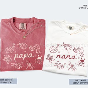 Op de afbeelding: Twee T-shirts, &eacute;&eacute;n karmozijnrood en &eacute;&eacute;n wit, met insecten- en bloemmotieven. Het karmozijnrode shirt heeft het woord "papa" en het witte shirt heeft het woord "nana". Beide shirts hebben een Comfort Colors-label en een ketting.