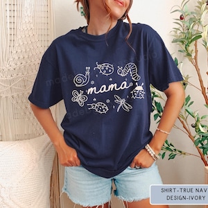 Op de afbeelding: Marineblauw T-shirt met het woord "mama" en insectenillustraties in wit. Het shirt heeft een ronde hals en korte mouwen. De tekst "SHIRT-TRUE NAVY DESIGN-IVORY" staat in een rechthoek onderaan.