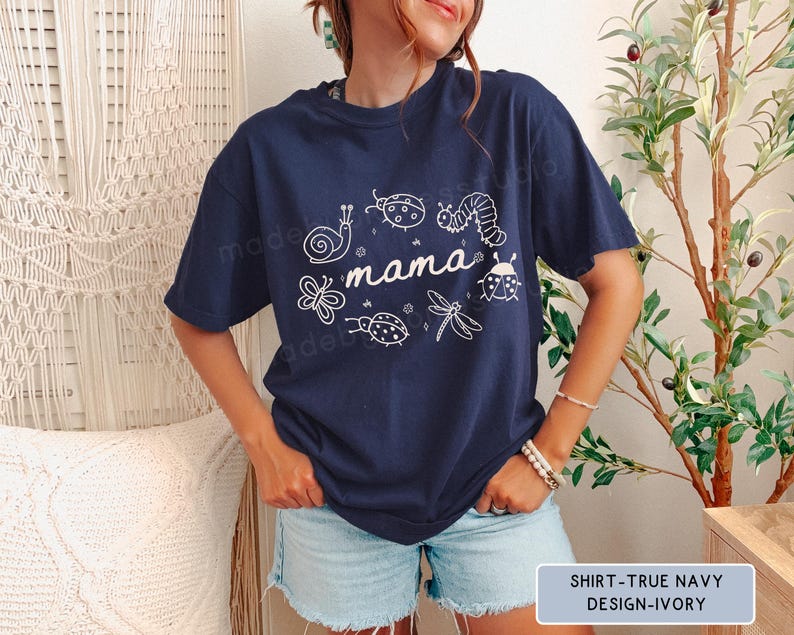 Op de afbeelding: Marineblauw T-shirt met het woord "mama" en insectenillustraties in wit. Het shirt heeft een ronde hals en korte mouwen. De tekst "SHIRT-TRUE NAVY DESIGN-IVORY" staat in een rechthoek onderaan.