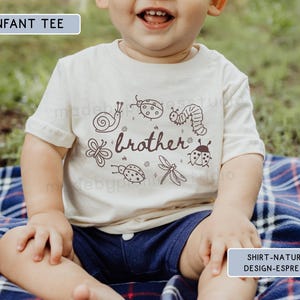 Op de afbeelding: Een cr&egrave;mekleurig baby-T-shirt met een bruine afbeelding van insecten en het woord "brother". Het ontwerp bevat een slak, lieveheersbeestjes, een rups, een vlinder, een libelle en andere insecten. Het kind draagt een marineblauwe korte broek en zit op een blauw geruite deken.