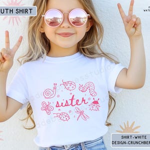 Op de afbeelding: Wit kinder-T-shirt met een roze afbeelding van insecten en het woord "sister". De persoon die het T-shirt draagt, draagt een roze zonnebril en maakt een vredesteken met haar vingers.